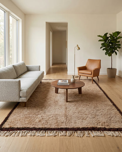 A plush tan bordered Beni Ourain Moroccan rug by NURA styled in a modern living room with a grey sofa and leather chair.