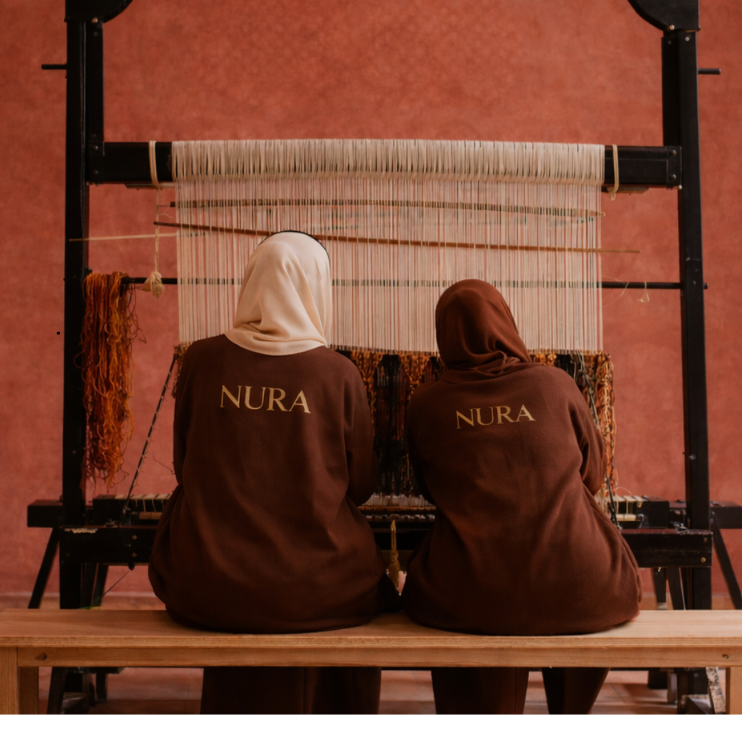 Two NURA artisans sitting at a traditional loom, handweaving a Moroccan wool rug and showing the craftsmanship behind each handmade piece.