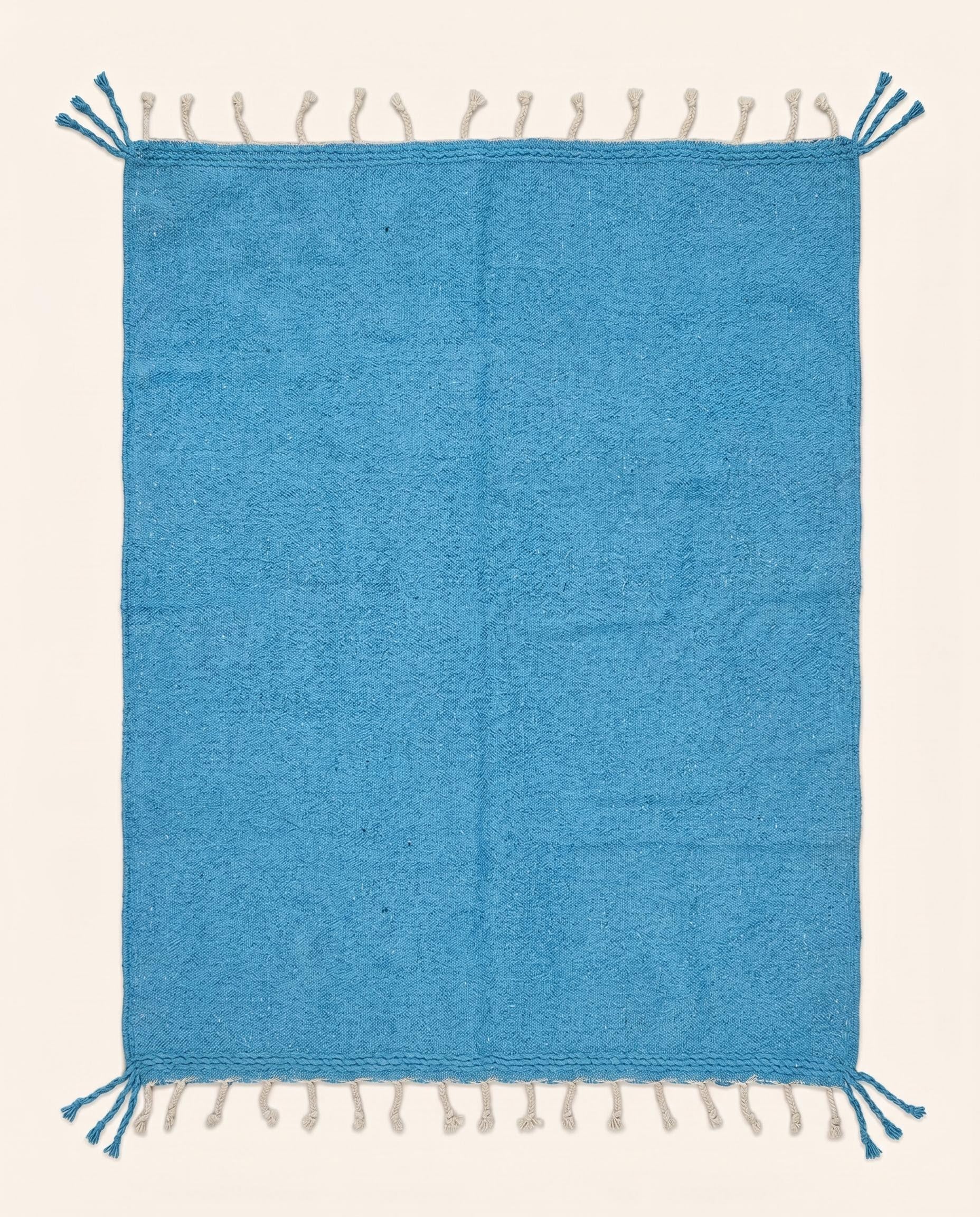 A vibrant solid azure blue Moroccan flatweave rug handwoven from natural wool, featuring braided fringes by NURA.