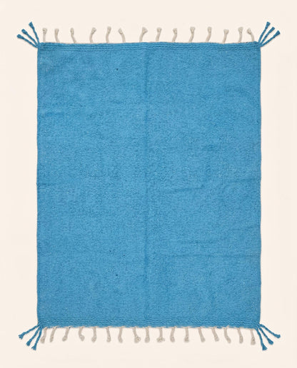 A vibrant solid azure blue Moroccan flatweave rug handwoven from natural wool, featuring braided fringes by NURA.